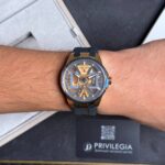 Wrist wearing a gold Ulysse Nardin skeleton watch with a blue strap, close-up view on forearm over a dark gift box.