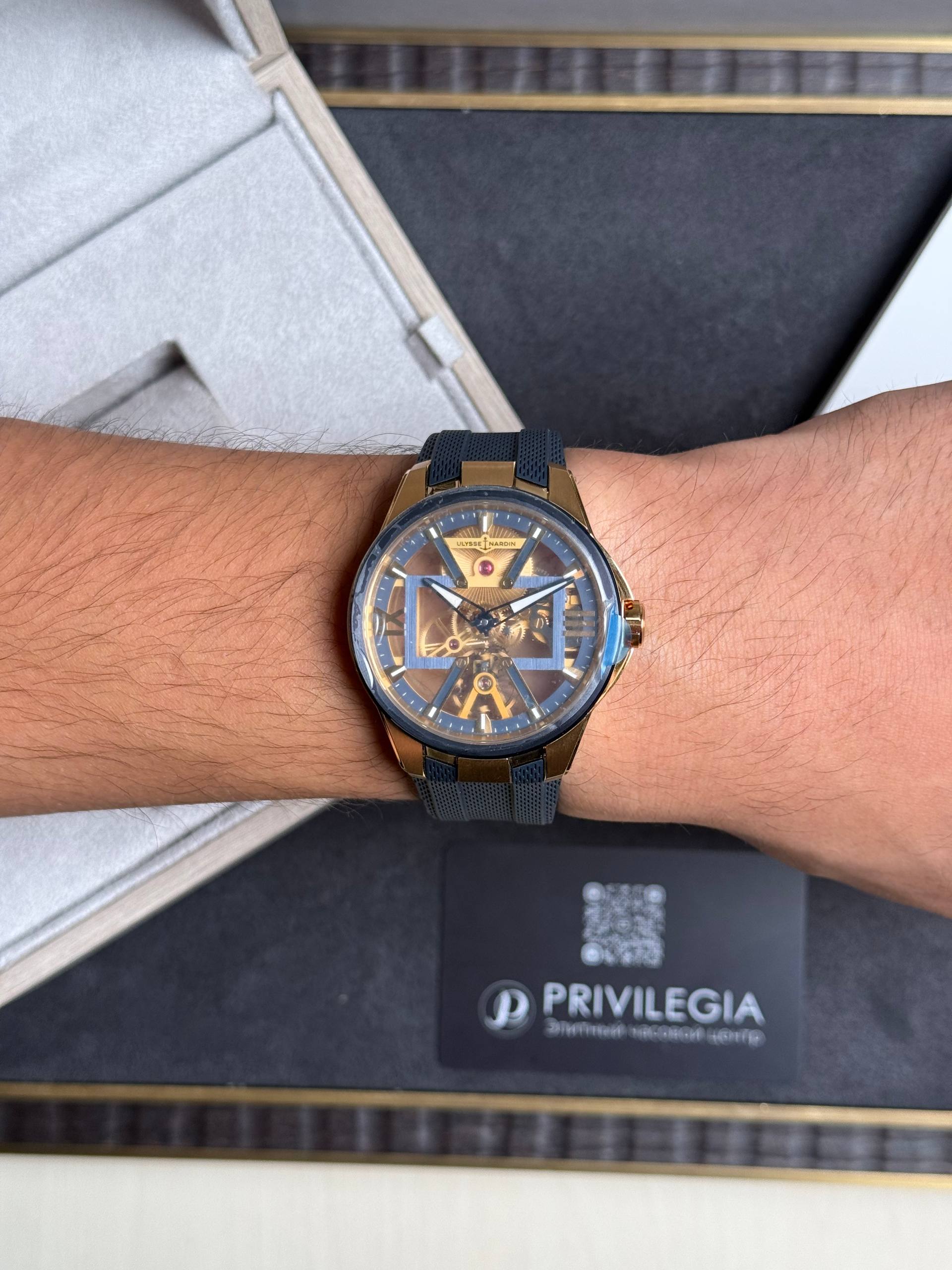Wrist wearing a gold Ulysse Nardin skeleton watch with a blue strap, close-up view on forearm over a dark gift box.