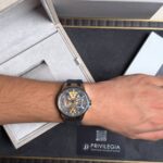 Wrist wearing a gold skeleton watch with a blue strap, resting next to an open jewelry box.