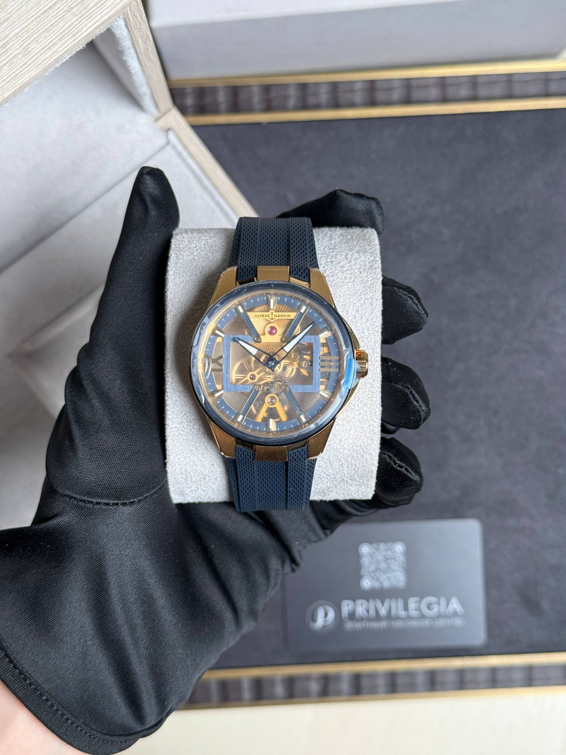 Skeleton gold-tone wristwatch with a blue perforated strap resting on a display cushion, Ulysse Nardin branding visible on the dial.