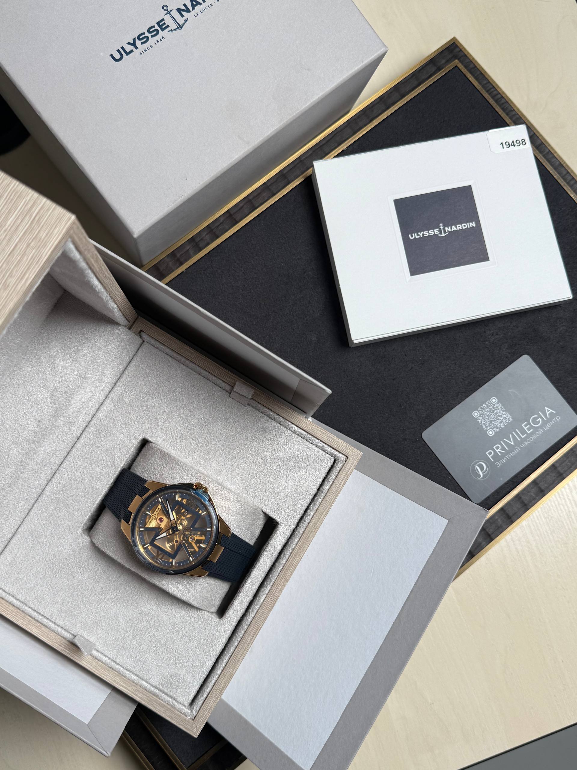 Luxurious wristwatch with a black strap displayed in an open gray box among other packaging and a Ulysse Nardin logo box in the background.