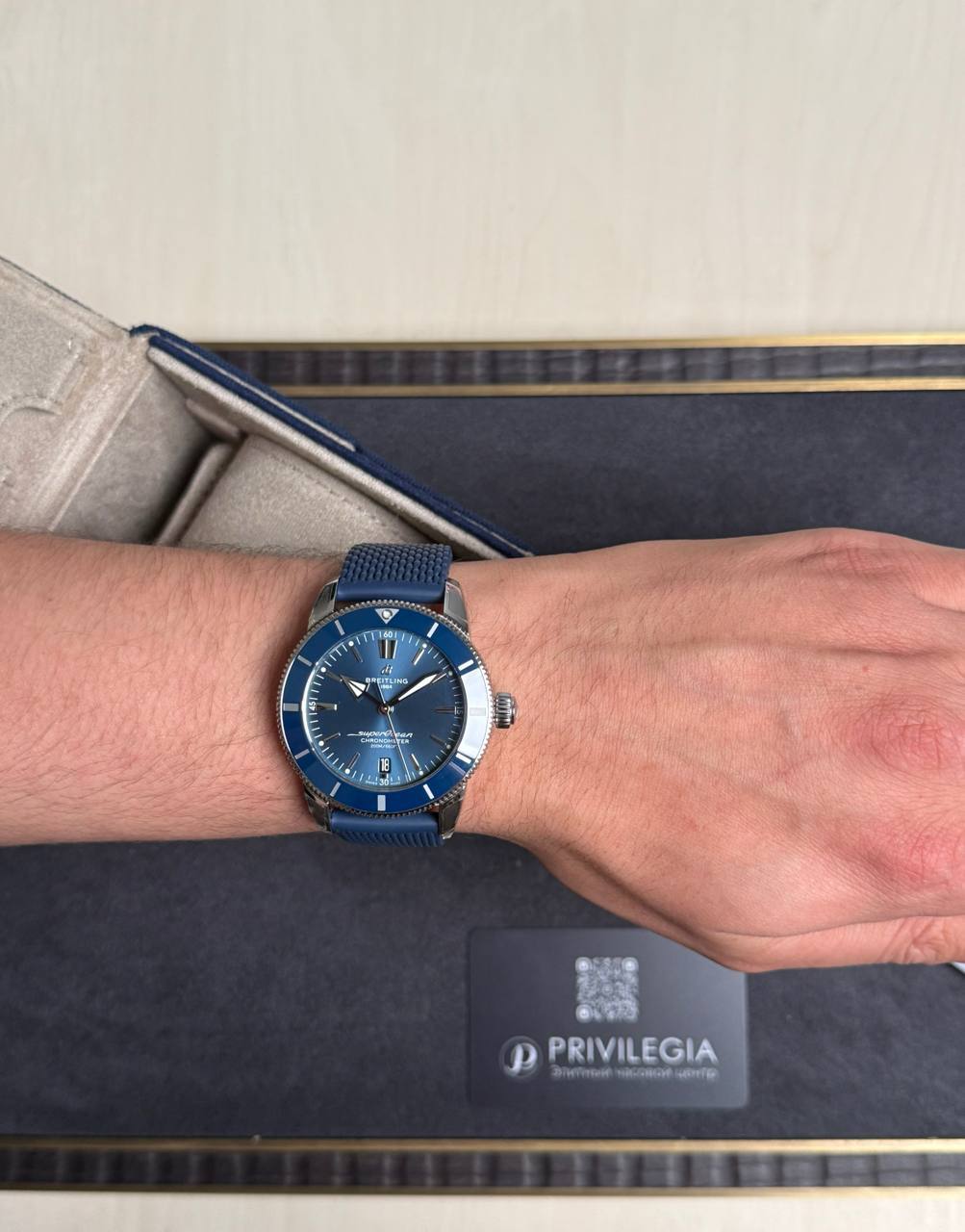 Blue Breitling wristwatch with a blue strap on a forearm, resting on a presentation box with a Privilegia card nearby.