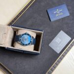 Blue Breitling wristwatch with a matching blue strap in its beige presentation box on a dark display surface, beside a gray Privlegia card and a blue booklet.