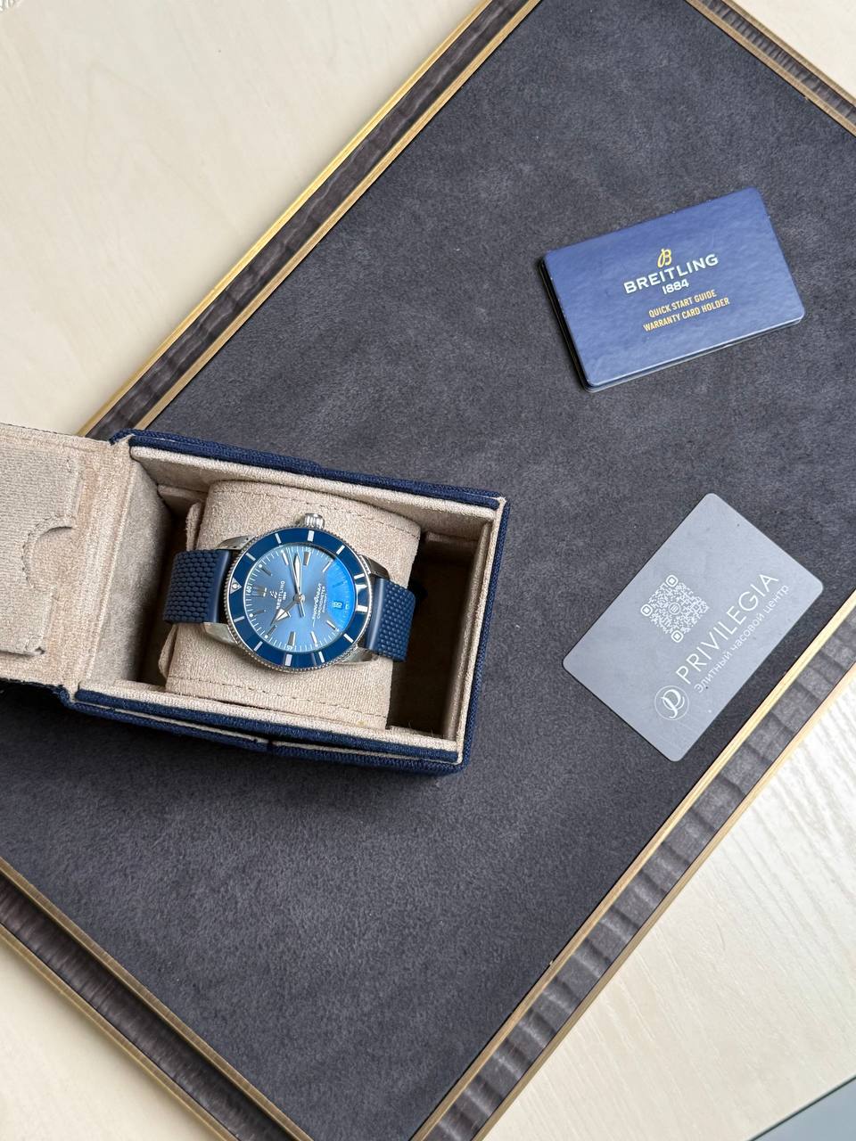 Blue Breitling wristwatch with a matching blue strap in its beige presentation box on a dark display surface, beside a gray Privlegia card and a blue booklet.