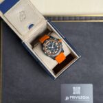 Sporty wristwatch with an orange strap in an open beige-lined box on a dark surface, Privilegia card nearby.