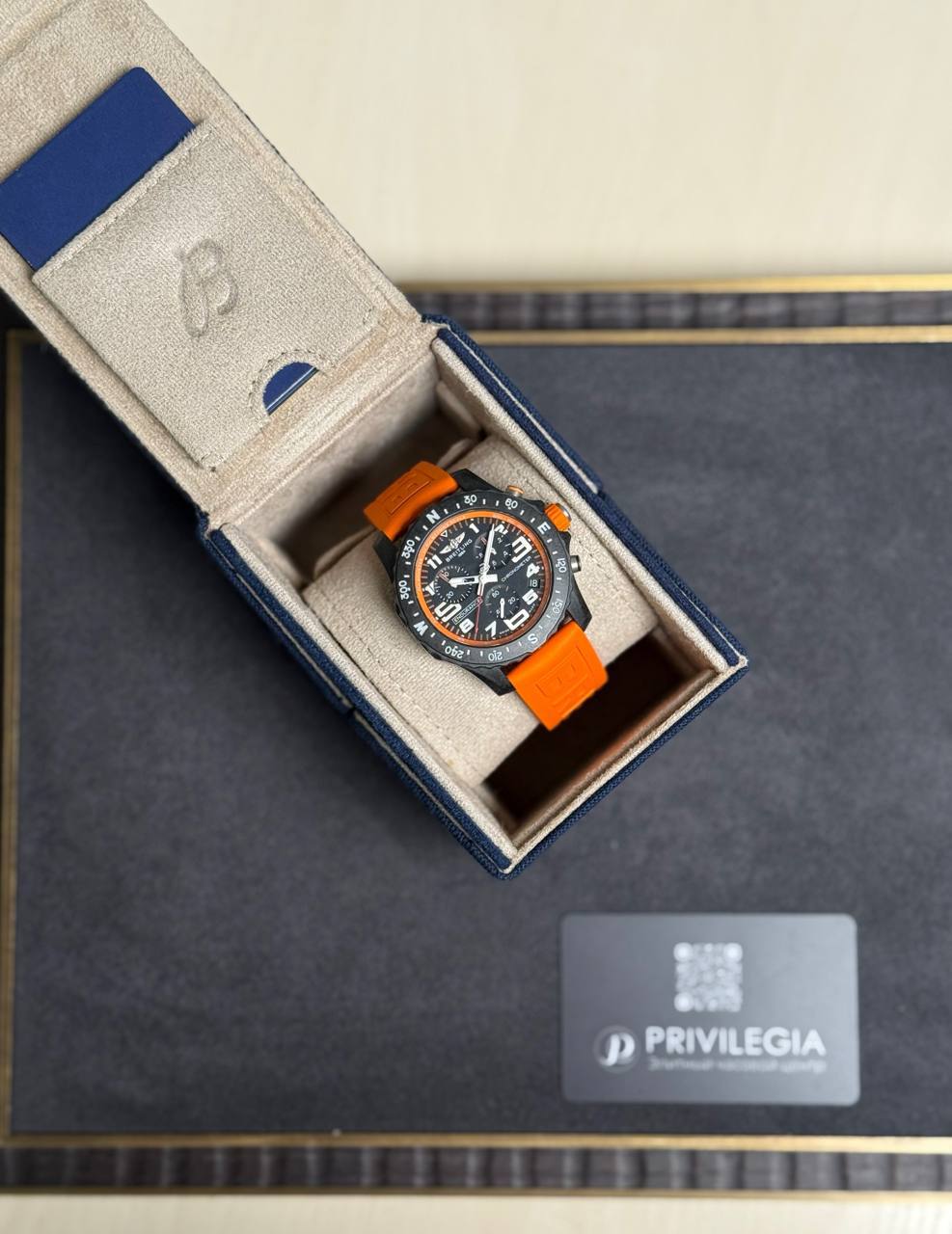 Sporty wristwatch with an orange strap in an open beige-lined box on a dark surface, Privilegia card nearby.