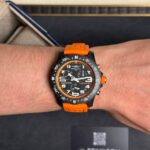 Wristwatch with black dial, orange bezel and orange strap on a forearm, Breitling Endurance in a display box in the background