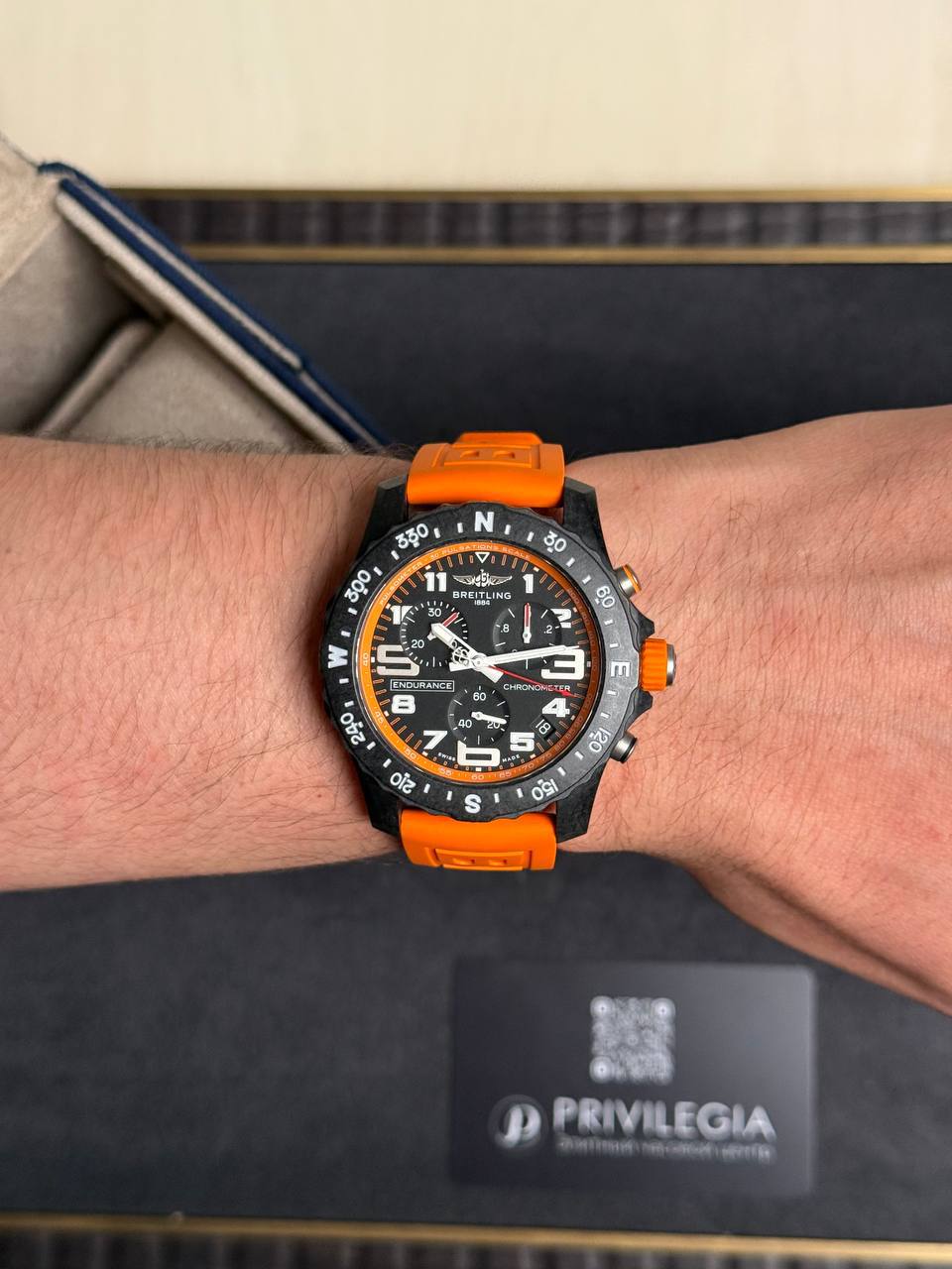 Wristwatch with black dial, orange bezel and orange strap on a forearm, Breitling Endurance in a display box in the background
