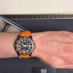 Wrist wearing a black Breitling chronograph with an orange strap, displayed in a presentation box.