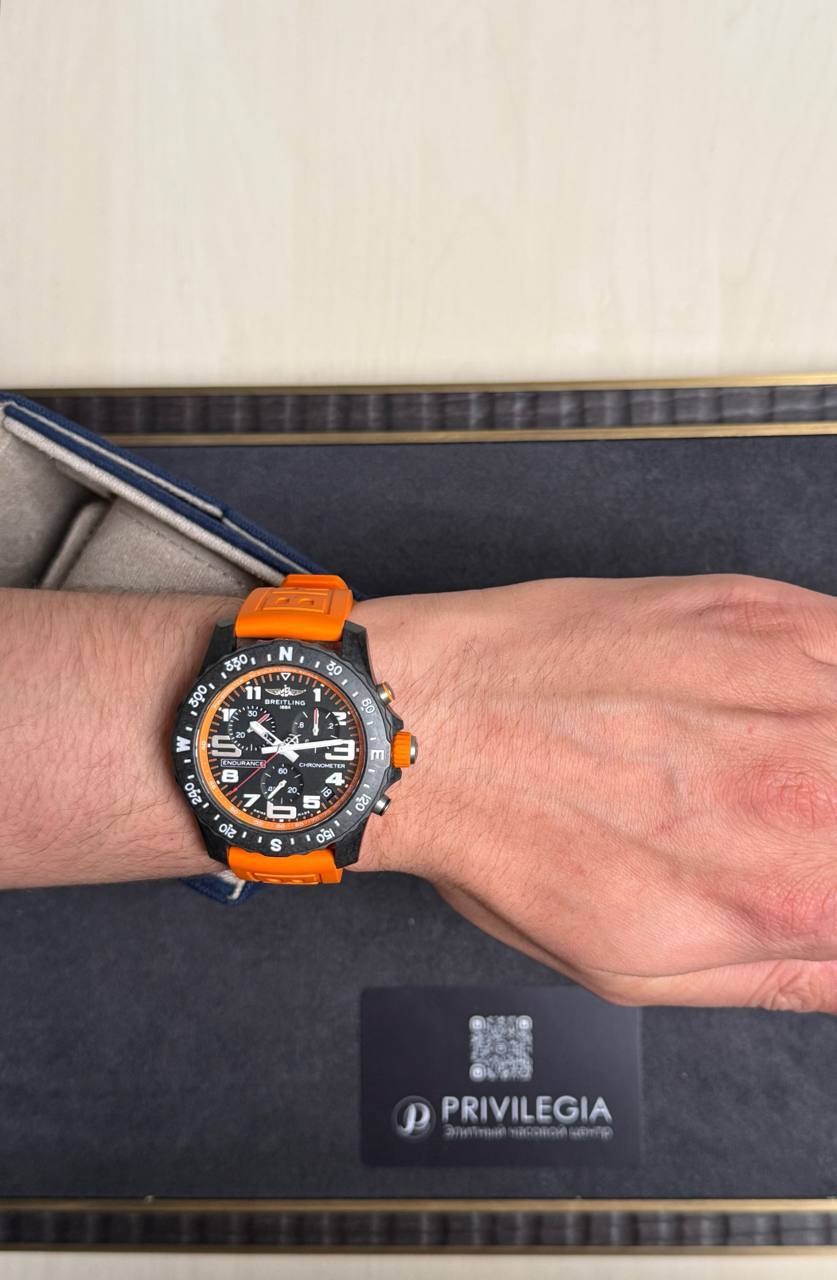 Wrist wearing a black Breitling chronograph with an orange strap, displayed in a presentation box.