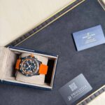 Breitling watch with an orange strap in its blue presentation box, warranty card nearby.