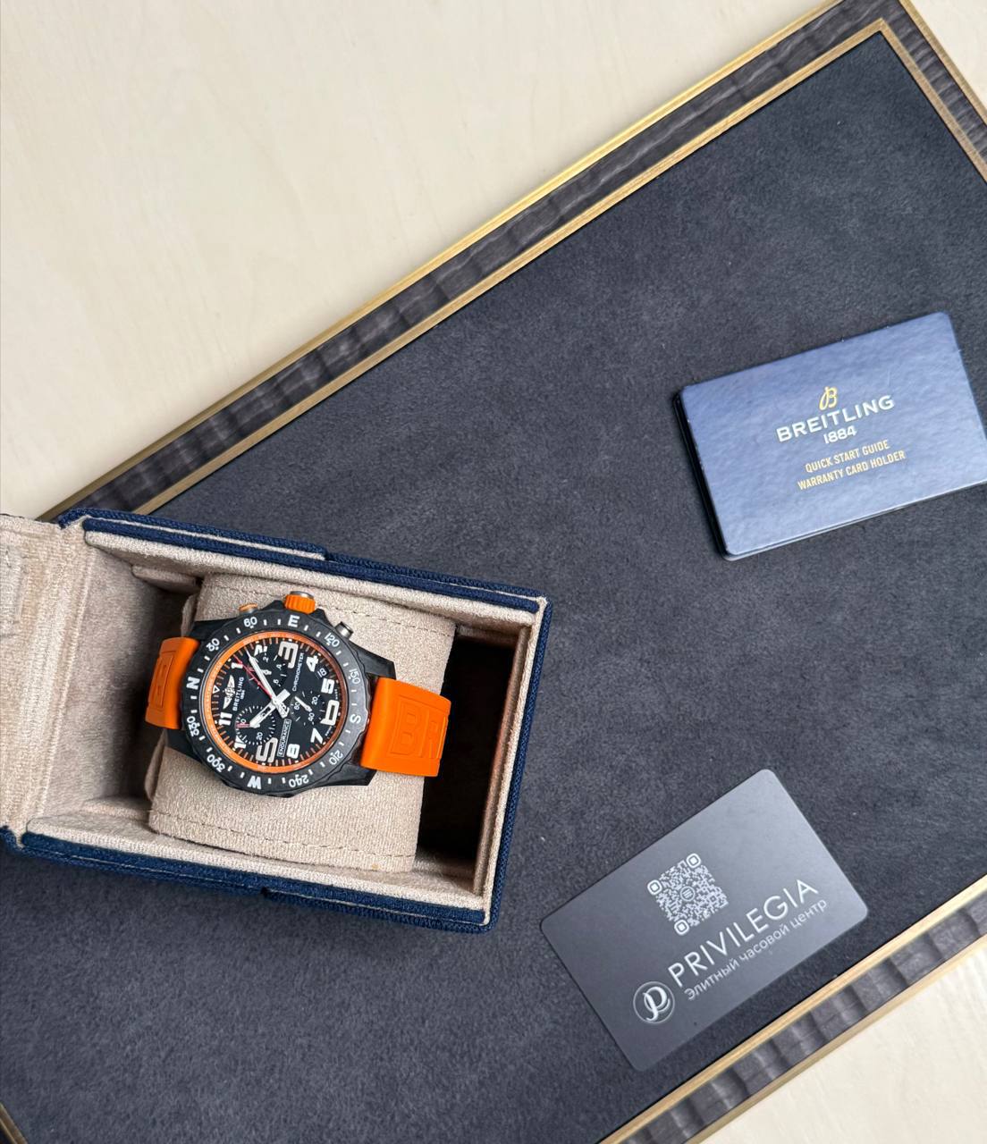 Breitling watch with an orange strap in its blue presentation box, warranty card nearby.