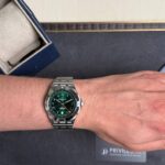 Wrist wearing a stainless steel Breitling watch with a green dial on a dark surface with a box nearby.