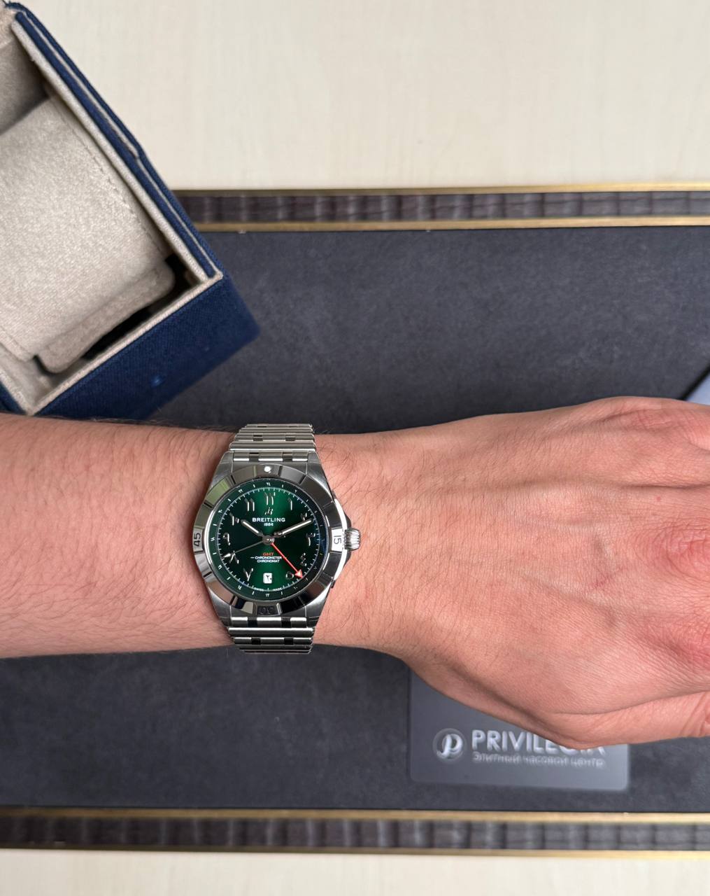 Wrist wearing a stainless steel Breitling watch with a green dial on a dark surface with a box nearby.