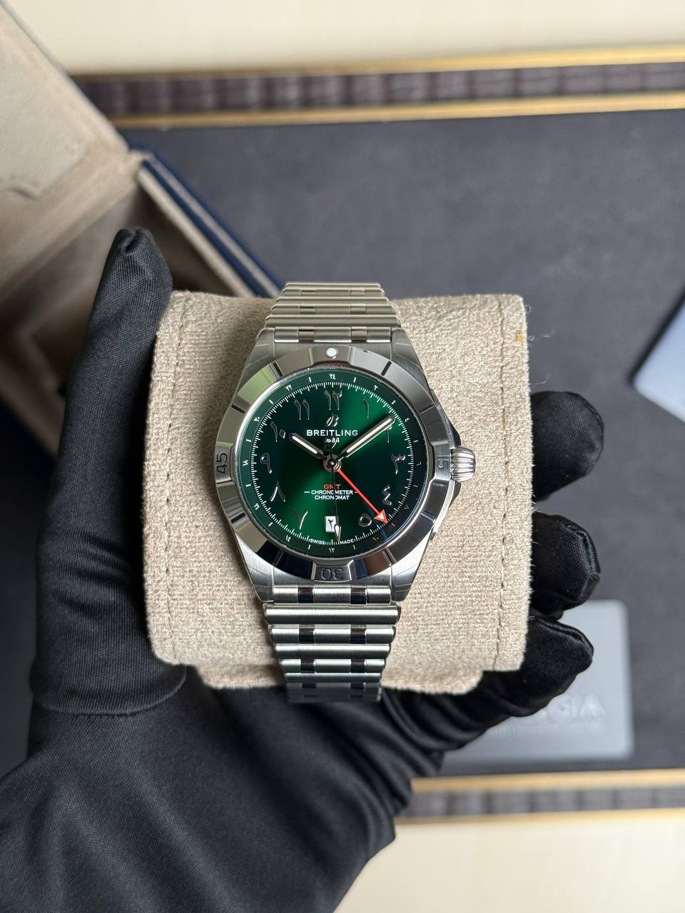 Breitling stainless steel wristwatch with green dial on a beige cushion, held by a gloved hand (dressy, luxury timepiece)