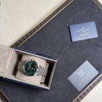 Breitling watch with green dial in an open blue presentation box, accompanied by warranty and Privilegia cards on a dark surface.