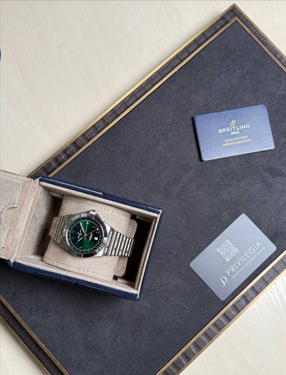 Breitling watch with green dial in an open blue presentation box, accompanied by warranty and Privilegia cards on a dark surface.