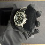 Black chronograph wristwatch with multiple subdials on a metal case, held in a gloved hand over a black velvet cushion.