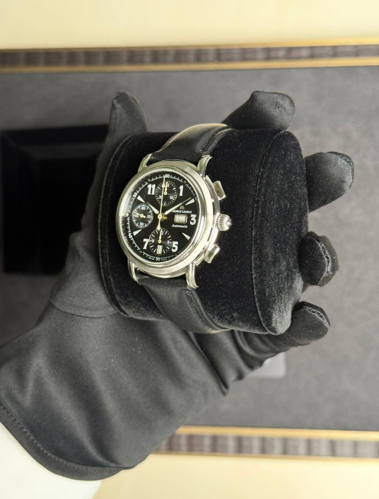Black chronograph wristwatch with multiple subdials on a metal case, held in a gloved hand over a black velvet cushion.