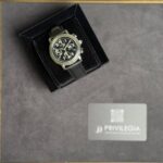 Black chronograph wristwatch with a leather strap in an open black display box on a gray surface, Privilegia card nearby