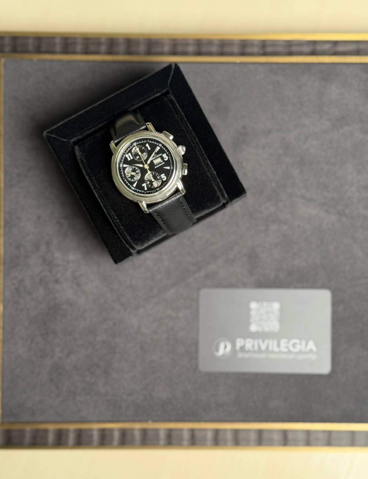 Black chronograph wristwatch with a leather strap in an open black display box on a gray surface, Privilegia card nearby