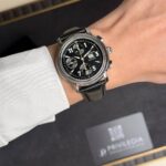 Close-up of a black-dial chronograph watch with silver case and black leather strap on a wrist, with a blurred box in the background.
