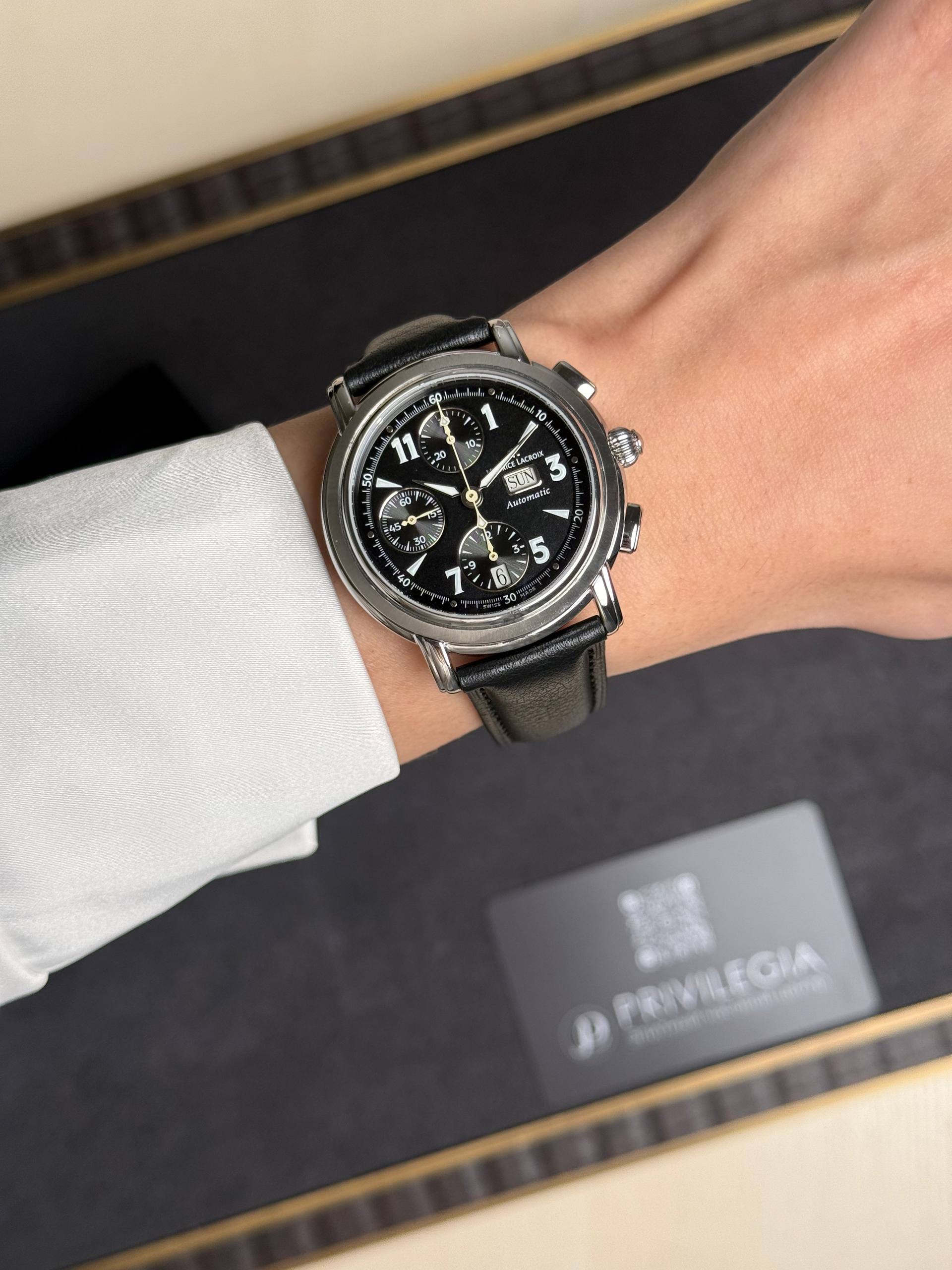 Close-up of a black-dial chronograph watch with silver case and black leather strap on a wrist, with a blurred box in the background.