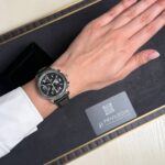 Left wrist wearing a black leather chronograph watch with white dial, resting on a dark display tray with a Privilegia card nearby.