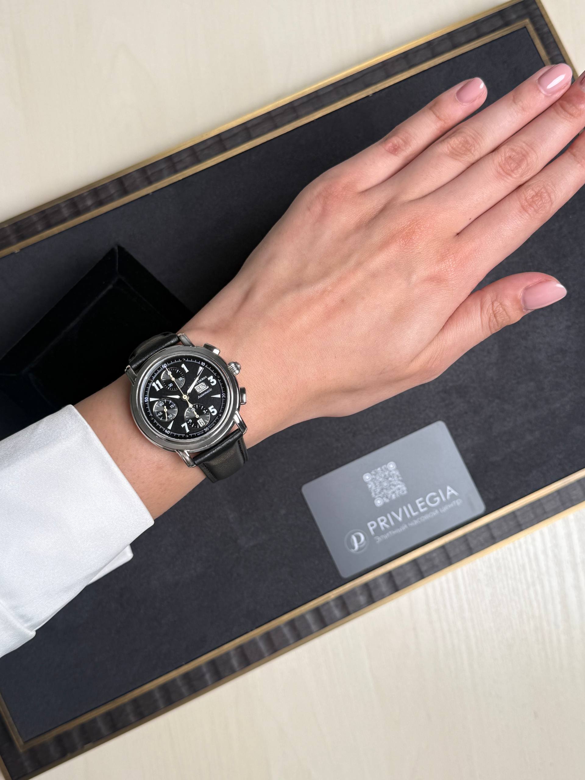 Left wrist wearing a black leather chronograph watch with white dial, resting on a dark display tray with a Privilegia card nearby.