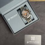 Tag Heuer watch in an open presentation box with a gray interior, metal bracelet and orange accents, beside a Privilegia card.