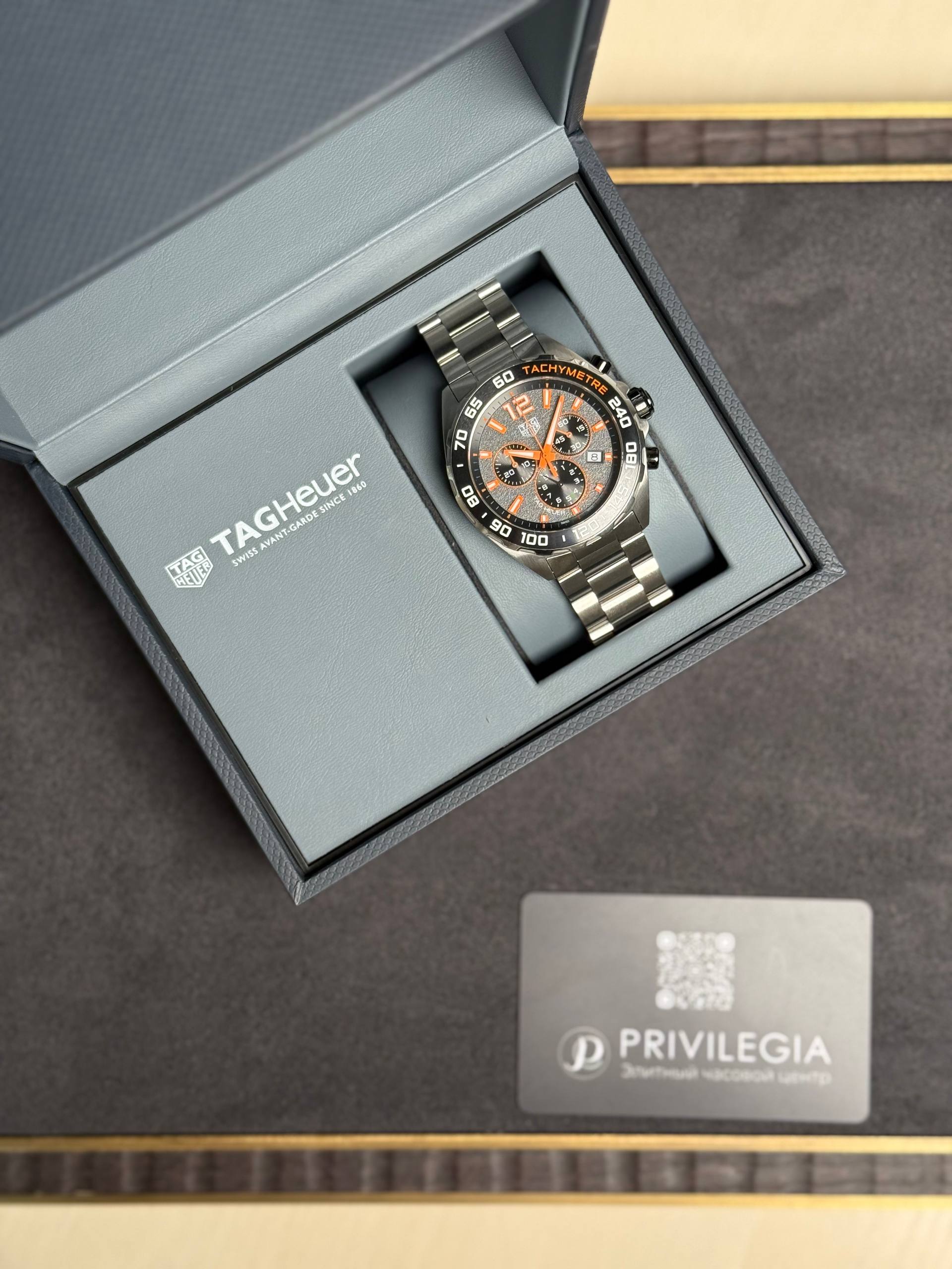 Tag Heuer watch in an open presentation box with a gray interior, metal bracelet and orange accents, beside a Privilegia card.