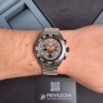 Wristwatch with orange accents and tachymeter bezel on a bare forearm, metal bracelet visible.