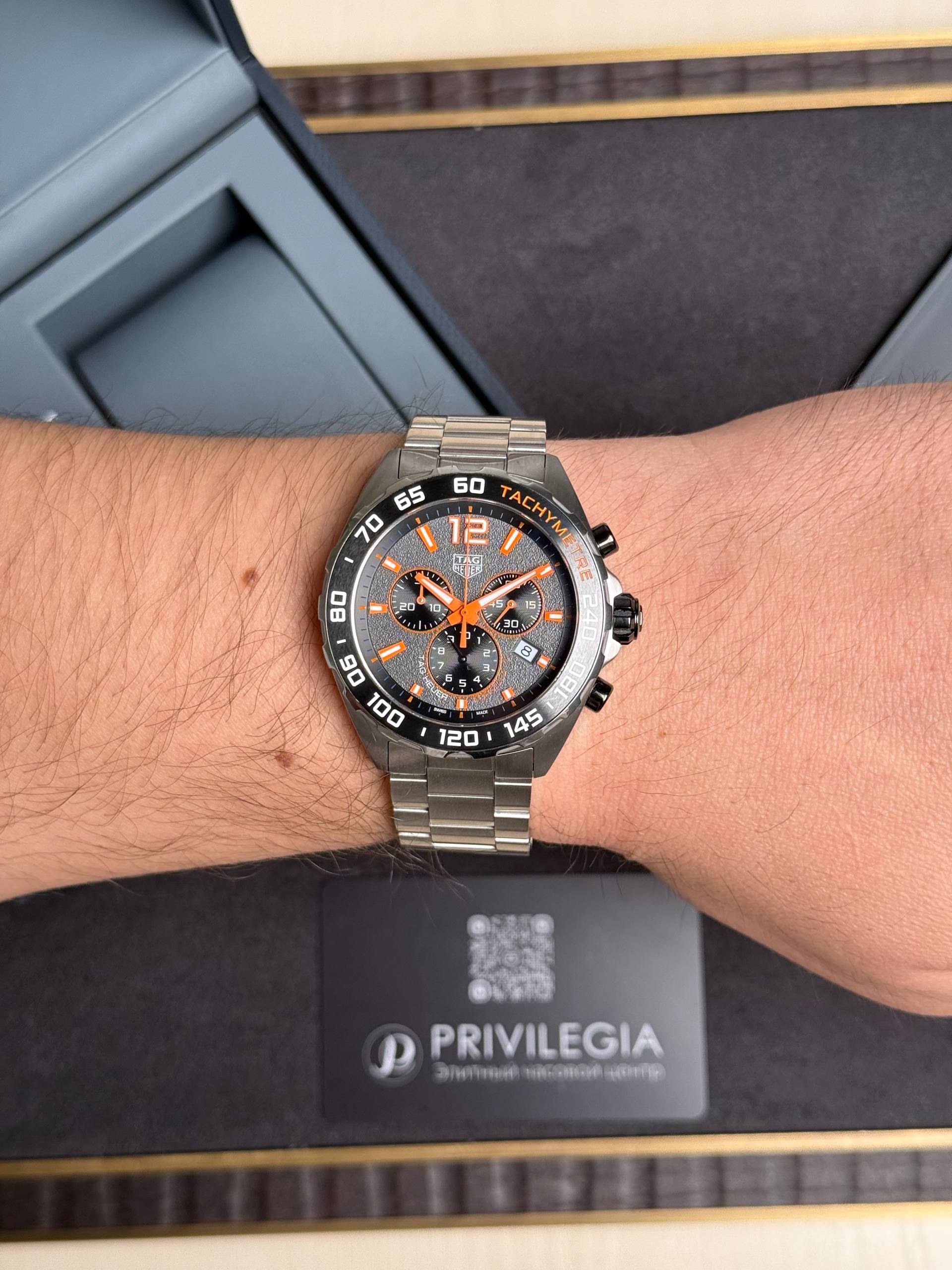 Wristwatch with orange accents and tachymeter bezel on a bare forearm, metal bracelet visible.