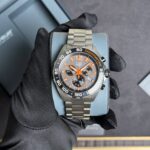 Stainless steel TAG Heuer chronograph with orange accents on a blue display cushion, held by a gloved hand.