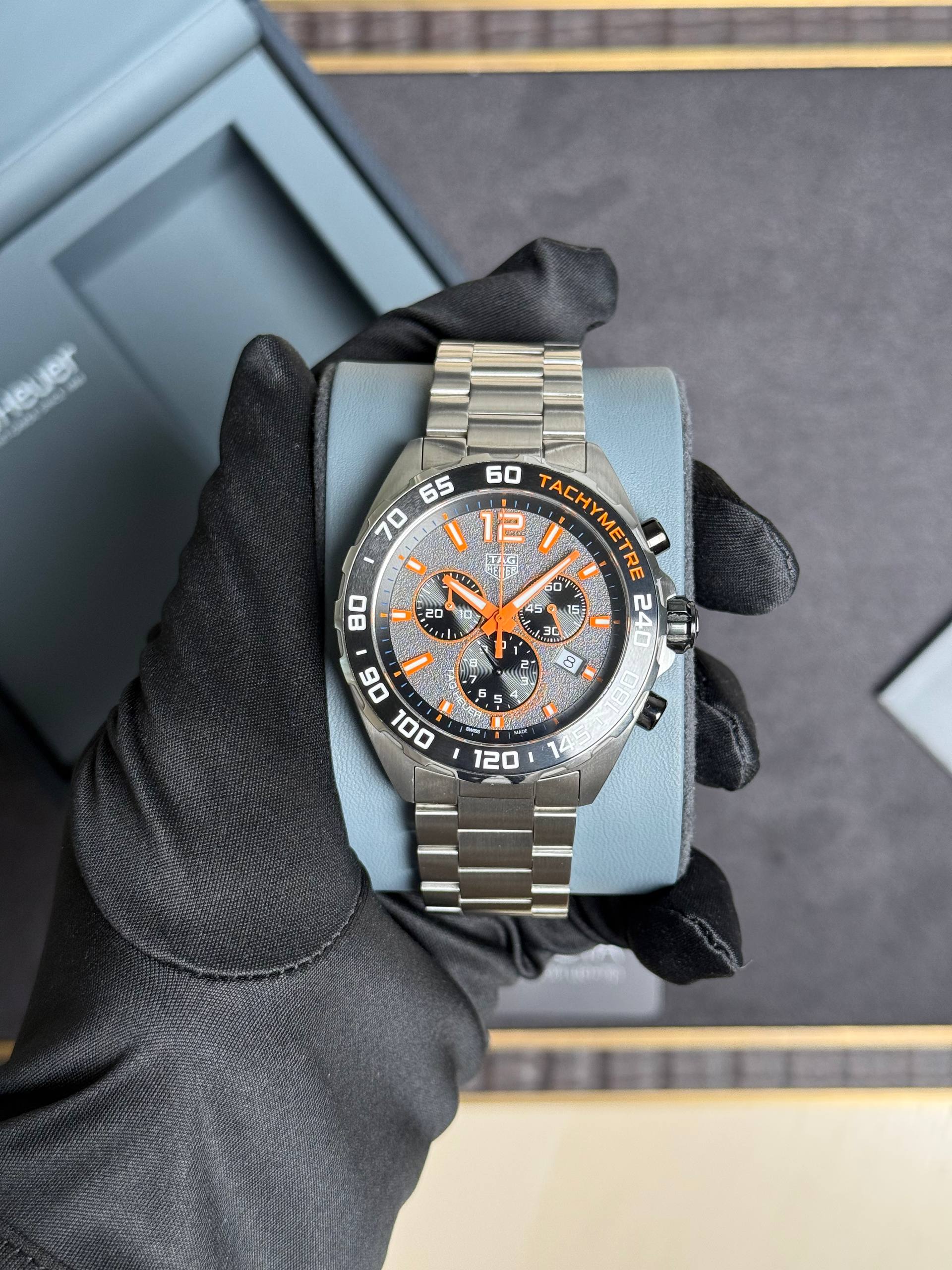 Stainless steel TAG Heuer chronograph with orange accents on a blue display cushion, held by a gloved hand.