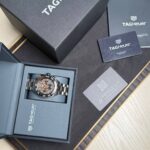 TAG Heuer watch in an open navy presentation box with accompanying guarantee card and booklet on a dark textured surface.