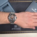 Close-up of a man's wrist wearing a silver TAG Heuer chronograph with orange accents, resting on a black box labeled 'Privilege'