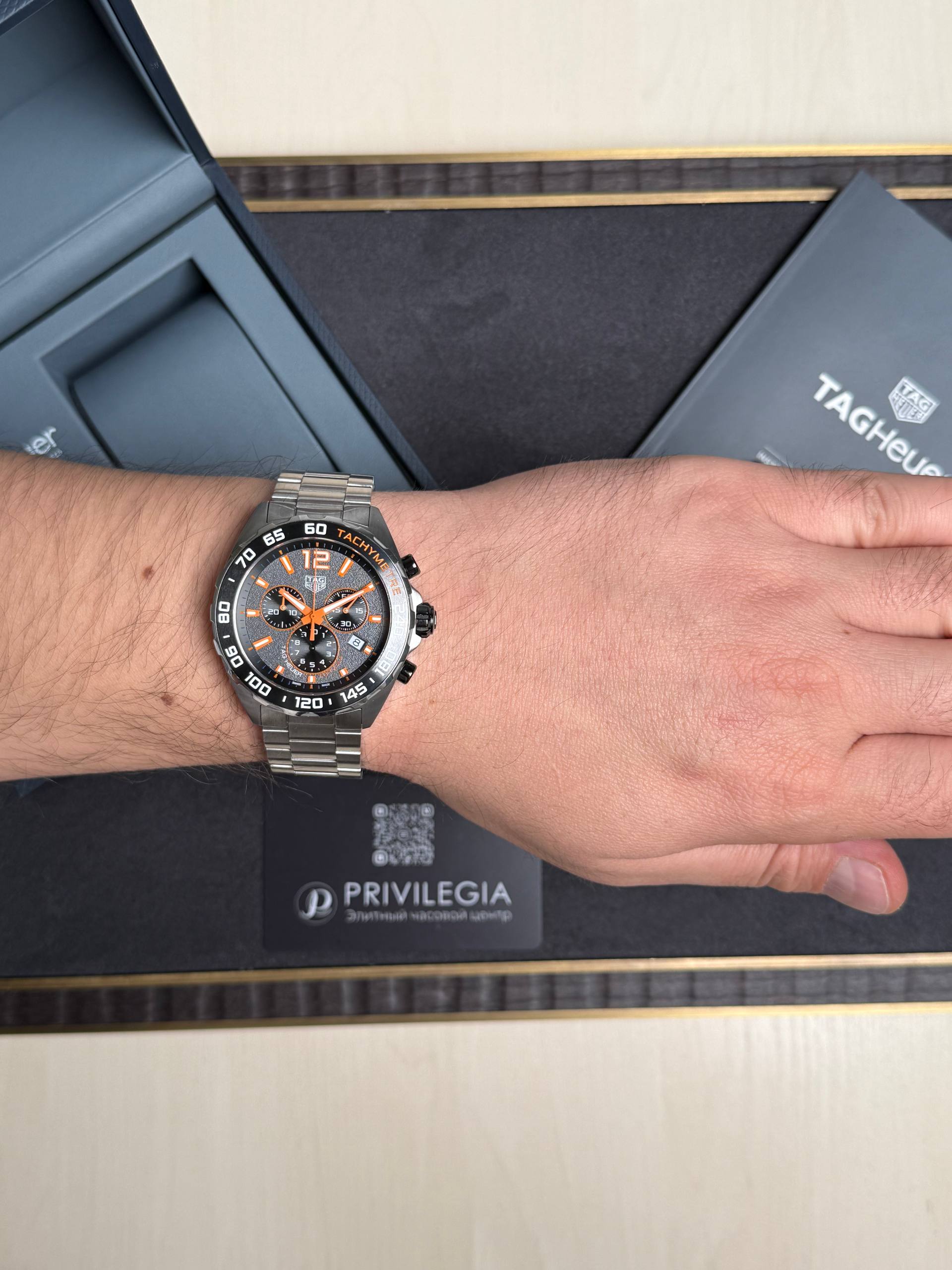 Close-up of a man's wrist wearing a silver TAG Heuer chronograph with orange accents, resting on a black box labeled 'Privilege'