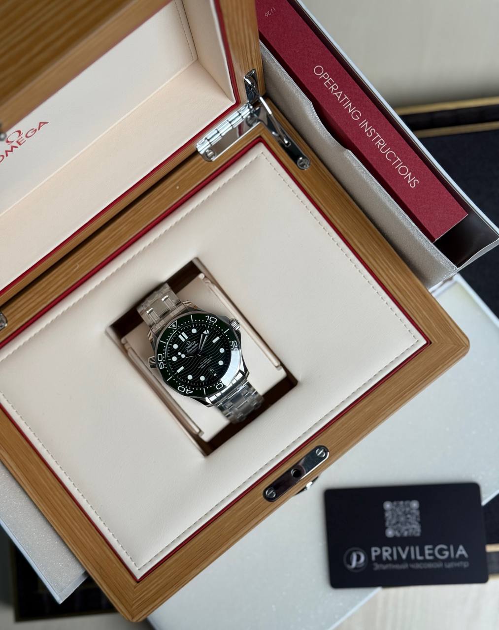 Omega diver-style stainless steel watch displayed in an opened wooden presentation box with a white interior and red trim, instruction booklet nearby