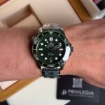 Wrist wearing a green-bezel Omega Seamaster diver's watch with a stainless steel bracelet.