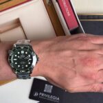 Omega Seamaster diver’s watch on a man's left wrist with a stainless steel bracelet and black dial featuring white indices and a date window at 6 o'clock as background shows books and a Privilegia card.