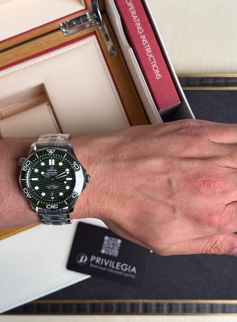 Omega Seamaster diver’s watch on a man's left wrist with a stainless steel bracelet and black dial featuring white indices and a date window at 6 o'clock as background shows books and a Privilegia card.