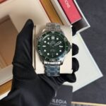 Close-up of a stainless steel Omega Seamaster watch with a green bezel and black dial, held by a gloved hand in a display box.