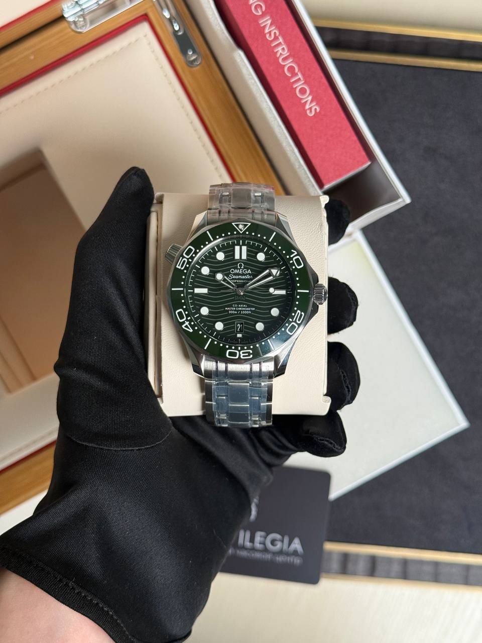 Close-up of a stainless steel Omega Seamaster watch with a green bezel and black dial, held by a gloved hand in a display box.