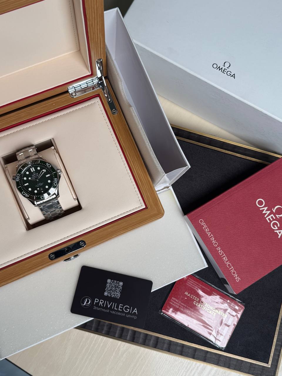 Omega stainless steel wristwatch in an open beige presentation box with red trimming, surrounded by Omega manuals and a Privilega card
