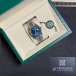 Rolex watch with a blue dial in an open green jewelry box, stainless steel bracelet, green seal tag nearby.