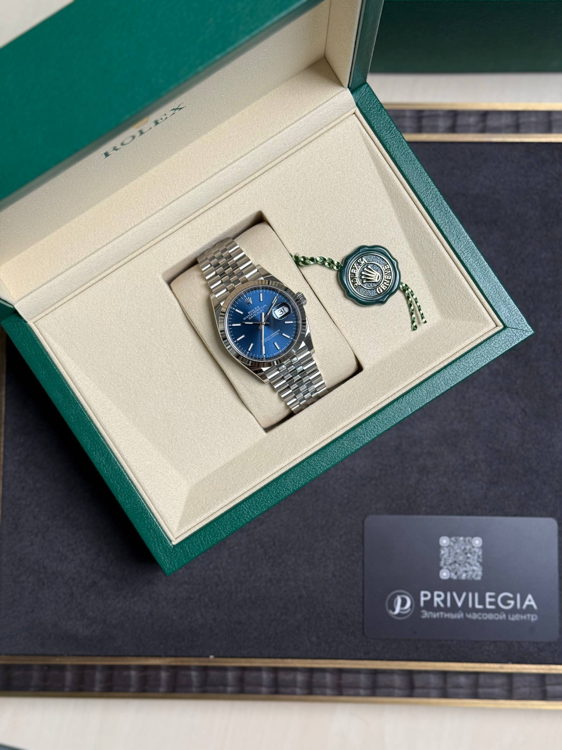 Rolex watch with a blue dial in an open green jewelry box, stainless steel bracelet, green seal tag nearby.