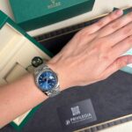 Wrist wearing a stainless steel Rolex watch with a blue dial, resting on an open green Rolex box.