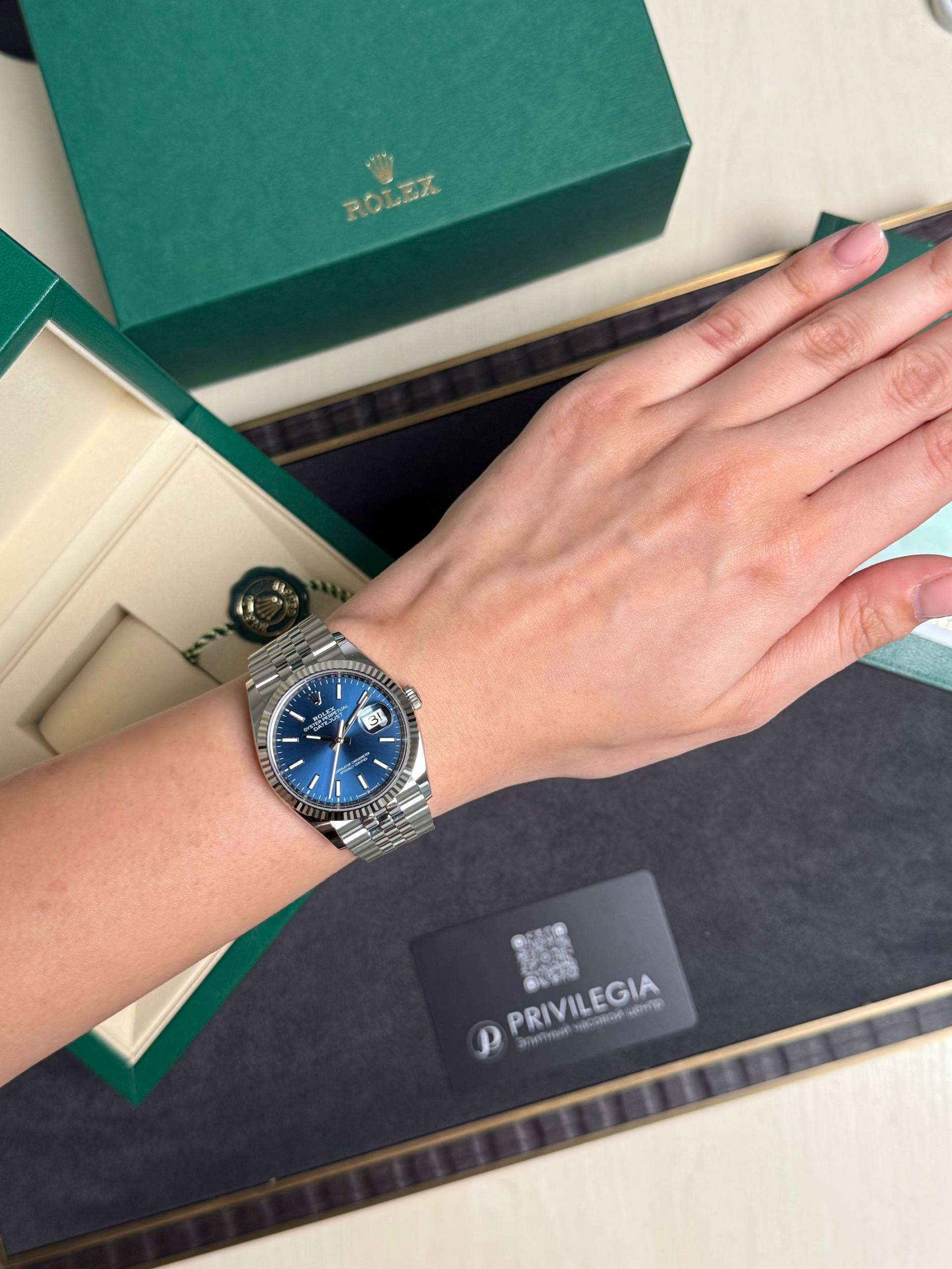 Wrist wearing a stainless steel Rolex watch with a blue dial, resting on an open green Rolex box.