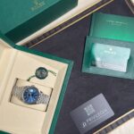 Green Rolex presentation box opened to reveal a silver bracelet watch with a blue dial and accompanying paperwork.